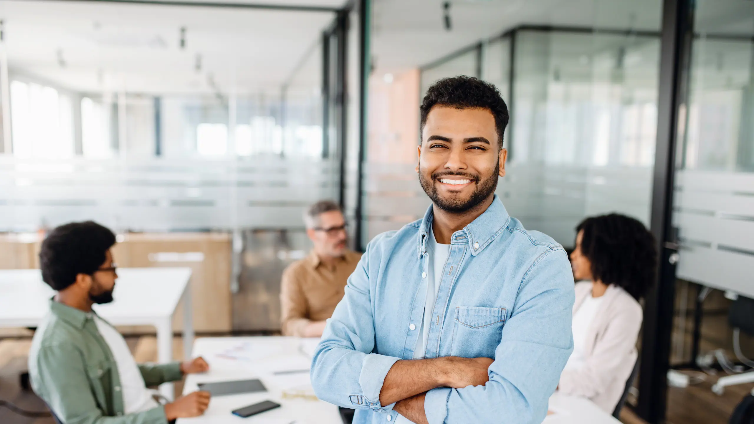 A confident young Indian male office employee in blue stands with his arms crossed in the foreground, with his team in discussion behind him, signaling a modern and proactive work environment