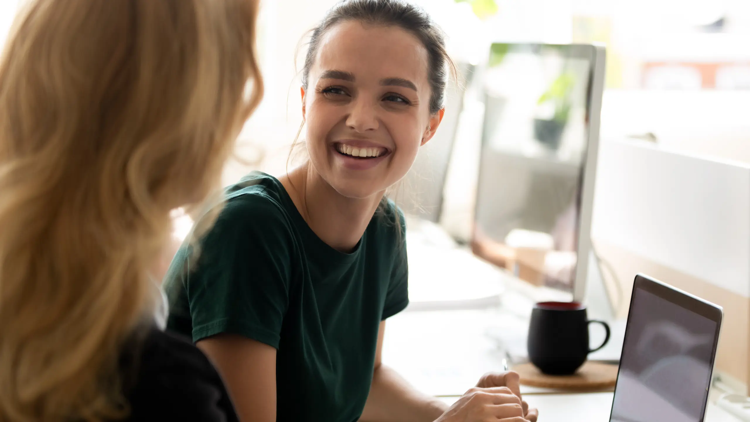 A marketer working with her colleague, collaborates on a laptop in a brightened room. They smile at each other.