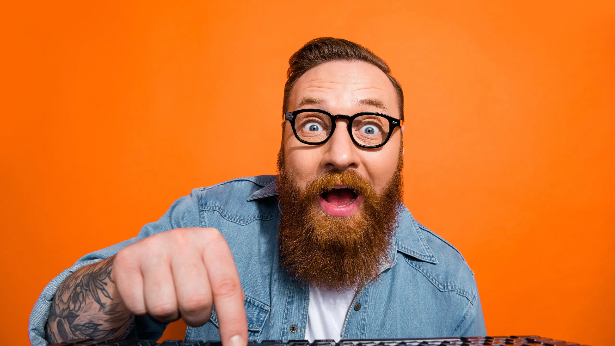 Image: A millennial man with a bushy beard and tattoos enthusiastically presses a button on his keyboard looking towards his screen with excitement.